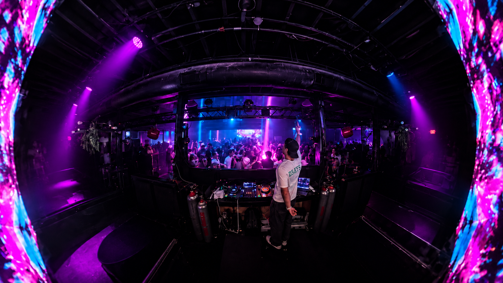 View from behind the DJ booth toward a packed crowd under colorful stage lights